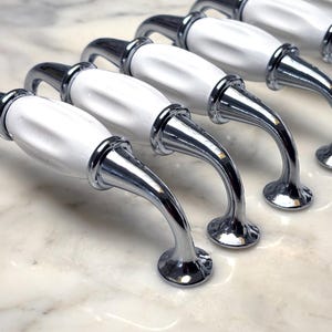 May include: A row of six cabinet pulls with white ceramic handles and polished chrome hardware. The handles have a curved design, and the pulls are arranged diagonally on a marble surface. The pulls are designed for furniture.
