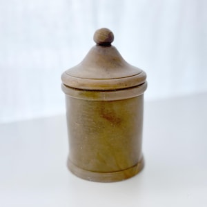 May include: A small, cylindrical container with a lid. The container is a muted brown color and has a rounded top with a small knob. The lid fits snugly on the container, creating a seamless look. The container is likely made of ceramic or stone.