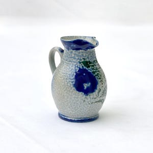 Peut inclure: Petite cruche en céramique avec une anse et un bec verseur. La cruche est principalement grise avec des accents bleus et verts. Un grand motif floral bleu orne le devant. Le bord et la base sont également bleus. Fond blanc.