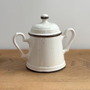 Peut inclure: Un sucrier en céramique blanc avec un couvercle et deux poignées. Le bol a une finition mouchetée avec une bordure marron foncé autour de la base, du couvercle et des bords des poignées. Le couvercle a un petit bouton sur le dessus.