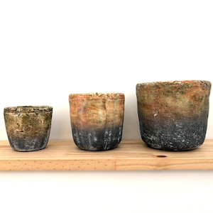 May include: Three ceramic pots of different sizes, all with a dark grey glaze and a light brown and green speckled pattern. The pots are arranged in a row on a wooden shelf.