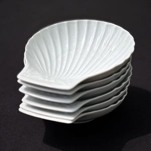 Peut inclure: Une pile de six petites assiettes blanches en forme de coquillage. Chaque assiette présente un motif nervuré en éventail partant du centre. Les assiettes sont empilées sur une surface sombre, la première étant bien nette.