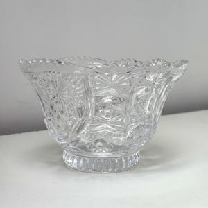 May include: A clear, cut-glass bowl with a scalloped rim and a pedestal base. The bowl features intricate geometric and floral patterns, reflecting light. The design includes starburst and fan-like shapes, creating a vintage aesthetic.