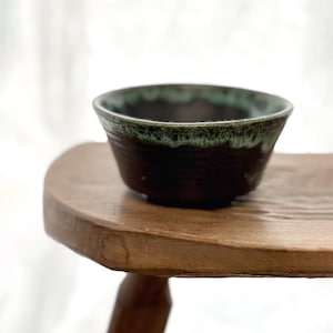 Peut inclure: Un bol en céramique fait à la main avec un extérieur brun foncé et un bord vert. Le bol est posé sur un tabouret en bois. L'intérieur du bol est brun foncé. Le bord a un aspect texturé et irrégulier.