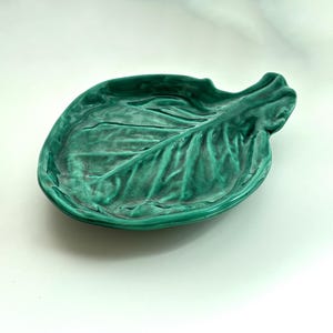 Puede incluir: Un plato de cerámica de color verde intenso y brillante con forma de hoja grande y texturizada. La superficie presenta venas en relieve y un contorno orgánico realista con un tallo prominente que se extiende por un lado, acabado con un esmalte liso.