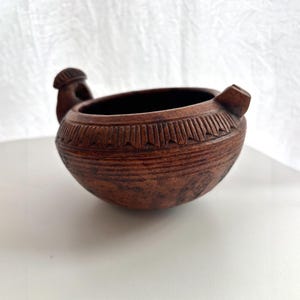 May include: A small, round, brown clay bowl with a textured surface. The rim features a carved geometric pattern of repeating triangles. It has two small, protruding handles on opposite sides, one shaped like a small knob and the other like a loop.