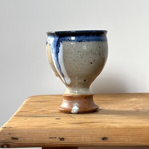 Peut inclure: Un gobelet en céramique fait à la main avec un corps beige moucheté, un bord bleu foncé et une base marron. Le gobelet a une forme unique avec un dessus évasé et une base en piédestal. Des coulures de glaçure blanche décorent le côté.