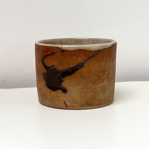 Puede incluir: Una taza cilíndrica de cerámica con exterior marrón y un diseño abstracto marrón oscuro. El borde es de color beige claro. La taza es probablemente hecha a mano y tiene una estética rústica.