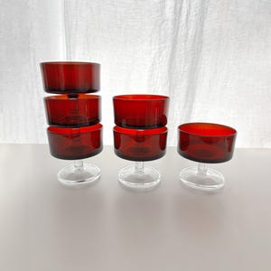 Puede incluir: Un conjunto de seis cuencos de postre de vidrio rojo rubí, de estilo vintage. Cada cuenco tiene forma cilíndrica y se asienta sobre una base transparente con pie. Los cuencos están dispuestos en grupo, mostrando su diseño y color.