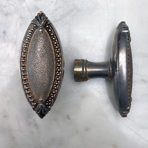 Peut inclure: Deux poignées de porte antiques argentées. L'une est ovale avec un centre texturé, une bordure perlée et des motifs de feuilles. L'autre a un design similaire et une tige de connexion. En métal.