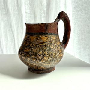 May include: A ceramic pitcher with a rounded body and a single handle. The surface features a dark red rim and a base, while the main body is decorated with intricate black geometric patterns, including triangles and cross-hatching on a tan background.
