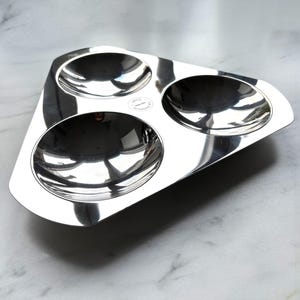Puede incluir: Una fuente brillante de acero inoxidable con tres compartimentos redondeados. La fuente tiene forma triangular con bordes redondeados y una pequeña etiqueta circular. La superficie reflectante crea un efecto de espejo.