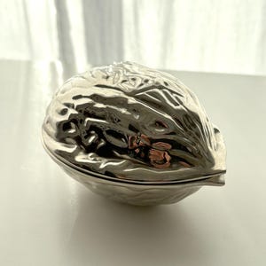 May include: A silver-coloured, walnut-shaped trinket box. The box is made of a shiny, reflective metal and has a detailed, textured surface that mimics the ridges of a walnut shell. The box is closed, with a visible seam where the lid meets the base.