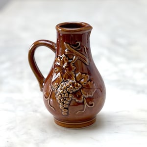 以下が含まれることがあります： ブドウの蔓のデザインが施された茶色の陶器のピッチャー。ピッチャーは持ち手と丸い本体があります。