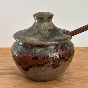 Könnte beinhalten: Ein kleiner, runder Honigtopf aus Keramik mit Deckel und einem Holzlöffel. Der Topf hat eine braune und graue Glasur mit einem gesprenkelten Muster.