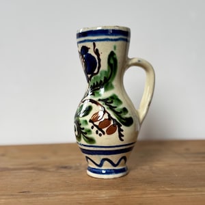 May include: A small, hand-painted ceramic pitcher with a cream-colored base. The pitcher features blue, green, and brown floral designs, a blue rim, and a small handle. The pitcher is approximately 5 inches tall.