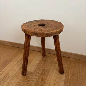 May include: A rustic wooden stool with a round top and three legs. The stool is made of natural wood and has a hole in the centre of the top.