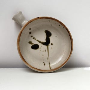 May include: A handmade ceramic bowl with a handle, featuring a cream-colored interior and a dark brown rim. The bowl is decorated with dark green and black paint splatters, adding a unique artistic touch. The handle is a light cream color.