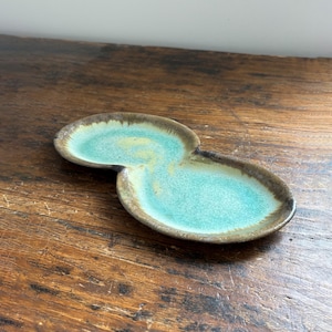 Peut inclure: Un plat de service en céramique fait à la main avec une forme double ovale unique. Le plat présente un intérieur turquoise et un bord marron. La surface a une finition brillante, et le plat est posé sur une surface en bois.