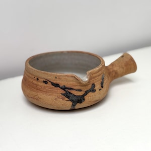 Puede incluir: Una cremera de cerámica hecha a mano con asa y boquilla. La cremera es de color marrón claro con salpicaduras de pintura negra oscura. El interior es de color gris claro.