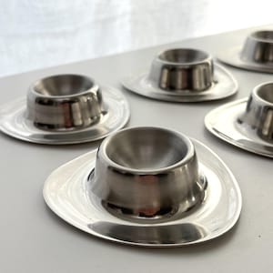 Peut inclure: Ensemble de six coquetiers en acier inoxydable. Chaque coquetier est composé d'une base circulaire avec un bord légèrement surélevé et d'une coupe centrale arrondie pour maintenir l'œuf. Les coquetiers sont disposés sur une surface gris clair.