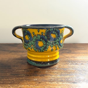Peut inclure: Un bol en céramique avec deux poignées, avec une base jaune vif et un intérieur bleu foncé. L'extérieur est orné d'un motif floral bleu et vert répété. Le bol a une base côtelée.