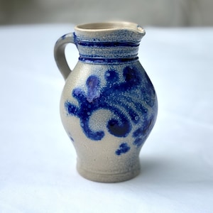 May include: A small stoneware jug with a handle and spout. The jug is mainly beige with blue floral designs and accents. The rim and handle are a darker blue. The jug is approximately 13 cm tall.