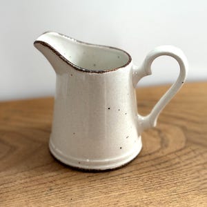 Op de afbeelding: Een kleine, crèmekleurige keramische creamer met een bruine rand en handvat. De creamer heeft een gespikkelde textuur en een gebogen tuit om gemakkelijk te schenken. De handgreep heeft een lusvorm. De creamer staat op een houten oppervlak.