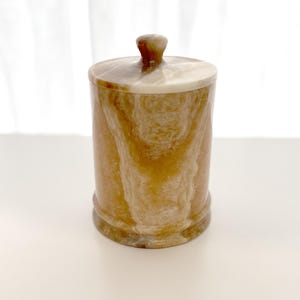 Puede incluir: Un tarro cilíndrico con tapa, hecho de piedra veteada. El tarro presenta una paleta de colores cálidos de beige, marrón y crema, con un pomo redondeado en la parte superior. Las vetas naturales de la piedra crean un patrón único.