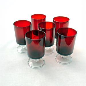 Peut inclure: Six verres en verre teinté rouge, avec des pieds et des bases transparents. Les verres ont une forme cylindrique courte et sont disposés sur une surface blanche. Les verres sont probablement destinés à servir des boissons.