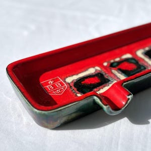 Könnte beinhalten: Ein rechteckiger Aschenbecher mit leuchtend roter Innenseite und dunkelgrüner Außenseite. Der Aschenbecher hat ein weißes Logo mit den Buchstaben "BB" und ein dekoratives Muster mit schwarzen und roten Akzenten. Eine kleine Kerbe ist sichtbar.