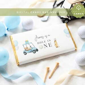Pode incluir: Uma embalagem retangular de barra de chocolate com fundo branco, um carrinho de golfe azul claro, uma bolsa de golfe e uma bola de golfe. O texto diz "James is a HOLE IN ONE" em letras pretas. O design inclui balões azul claro.
