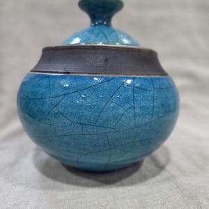 May include: A round ceramic jar with a lid, featuring a vibrant turquoise blue glaze with a prominent dark crackle pattern. The upper rim of the jar and the lid handle are finished in a contrasting matte dark charcoal gray, smooth texture.