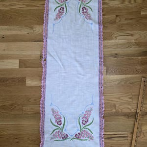 May include: A rectangular white linen table runner featuring embroidered pink and purple floral clusters at both ends, accented with light blue stitched lines and a pink crocheted lace border. A wooden ruler shows the length is 36 inches.