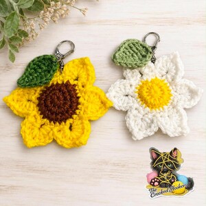 May include: Two crochet flower keychains with silver-tone lobster clasps. One is a yellow sunflower with a brown centre and a green leaf. The other is a white flower with a yellow centre and a green leaf. A sticker shows a black cat with yarn.