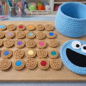 Op de afbeelding: Een verzameling handgemaakte gehaakte items, waaronder een blauwe mand, een Cookie Monster-onderzetter en talrijke gehaakte koekjes met kleurrijke centra. De items zijn gerangschikt op een houten oppervlak.