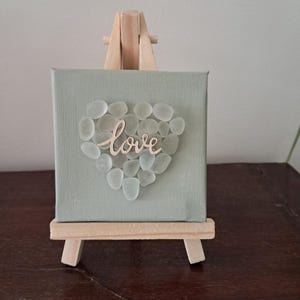 May include: A small, square canvas with a pale green background, featuring a heart of clear sea glass. The word "love" is written in cursive across the centre. The canvas is displayed on a miniature wooden easel.