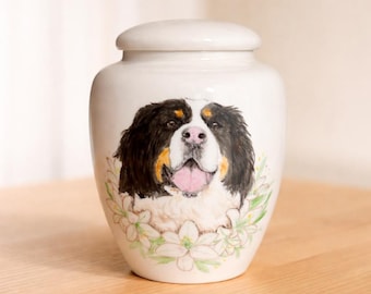 May include: A white ceramic urn featuring a hand-painted portrait of a Bernese Mountain Dog with black, white, and tan fur. The dog is depicted with its tongue out, surrounded by a decorative wreath of light green and white flowers.