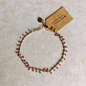 Può includere: Cavigliera dorata con perline smaltate colorate. La cavigliera presenta piccole perline pendenti in vari colori, tra cui rosso, rosa, blu e nero. Una piccola etichetta recita "Hand Made With Love Thank You".