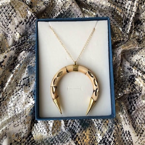 May include: A crescent-shaped pendant necklace featuring a curved, light-coloured material with inlaid metallic leaf-shaped accents and pointed gold-tone metal tips at both ends, suspended from a delicate gold-tone chain inside a blue box.