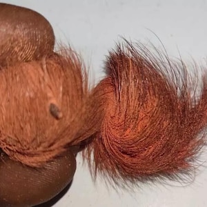 Può includere: Primo piano di un ciuffo di capelli rosso-brunastri, forse una parrucca o un posticcio. I capelli sono ricci e strutturati, con una piccola macchia scura visibile. Lo sfondo è una superficie bianca.