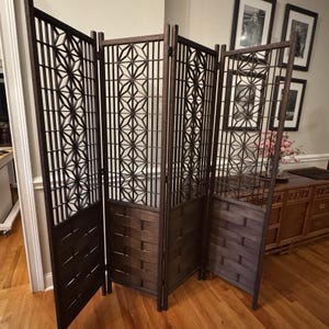 May include: A dark brown, four-panel room divider with intricate geometric cutouts. The lower sections feature a woven pattern. The divider is freestanding and adds a decorative element to any space. The panels are approximately 183 cm tall.