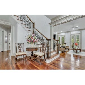 May include: An interior shot of a grand foyer with a staircase, hardwood floors, and elegant furnishings. Two ornate chairs flank a small round table with a floral arrangement. The staircase features decorative ironwork and a wooden handrail, leading to an upper level.
