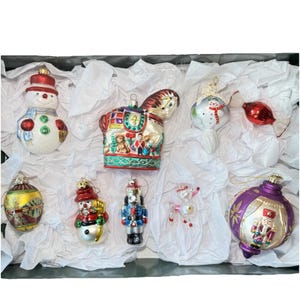 May include: A collection of Christmas ornaments in a black box, including a snowman, a horse, a snowman, a red ornament, a gold ornament, a snowman, a nutcracker, a reindeer, and a purple ornament. The ornaments are made of glass and have various colors and designs.