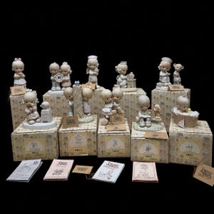Könnte beinhalten: Eine Sammlung von matten Porzellanfiguren von Precious Moments, die Kinder in verschiedenen Szenen darstellen, begleitet von ihren originalen Pappschachteln und Beipackzetteln. Die Figuren sind in sanften Pastelltönen bemalt.