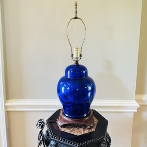 Op de afbeelding: Een levendige blauwe keramische tafellamp met een glanzende afwerking. De lamp heeft een klassieke gemberpotvorm, een messing hals en een decoratieve bekroning. Hij staat op een donkere, sierlijke tafel.