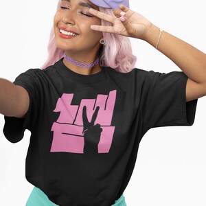 May include: A black t-shirt with a pink peace sign graphic and the text "I'm With The Band". The model is wearing a purple baseball cap and a light blue skirt.