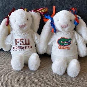 May include: Two white plush bunny toys wearing white hoodies. One hoodie displays "FSU FLORIDA STATE UNIVERSITY" in maroon, and the other features a green alligator and the word "Gators" in orange. Each bunny has colorful ribbons tied to its ears.
