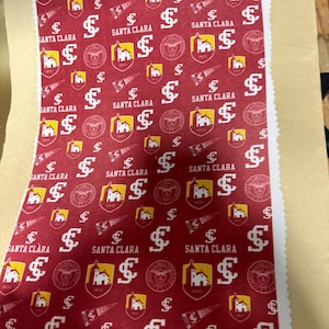 May include: A piece of fabric with a repeating pattern of Santa Clara University logos, including the letter "S", the university seal, and the words "Santa Clara". The background is a deep red color.