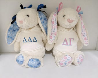Puede incluir: Dos conejos de peluche color crema con sudaderas blancas. El conejo izquierdo tiene orejas y pies con estampado azul y las letras griegas Delta Delta Delta, además de cintas azules. El derecho tiene estampado rosa y las letras Delta Gamma.